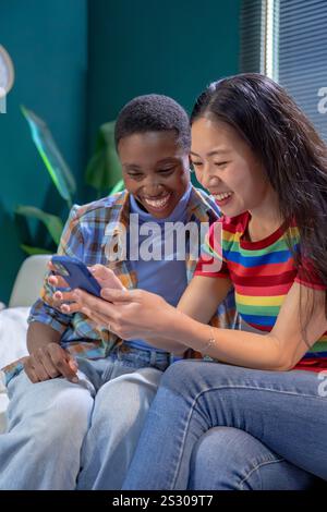 Donne nere e cinesi con smartphone in salotto. Amici, amicizia, tempo insieme Foto Stock