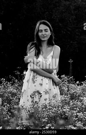 Una giovane donna in abito si abbraccia con gli occhi chiusi, in piedi in un campo a margherita Foto Stock