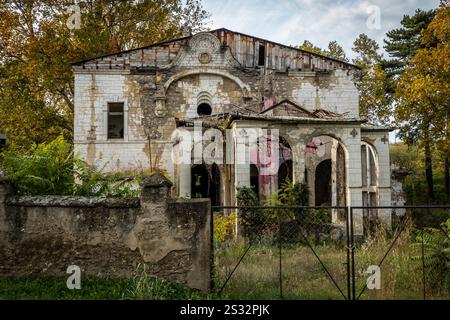 Una casa vecchia, trascurata e abbandonata, completa di una recinzione situata proprio di fronte ad essa, è attualmente circondata da alti alberi Foto Stock
