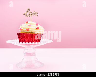 San Valentino. Un cupcake con crema e decorazione rossa con un segno di legno con la parola AMORE su sfondo rosa. Pasticceria. Foto Stock