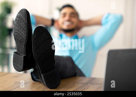Giovane dipendente arabo che si rilassa dopo un lavoro di successo, mettendo le gambe sul tavolo in ufficio, fuoco selettivo Foto Stock