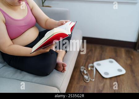 Donna in sovrappeso che legge un libro per perdere peso. Donna grassa preoccupata per la dieta del peso perdere stile di vita. Foto Stock