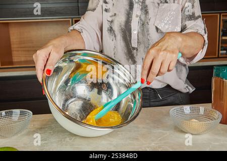 Una persona mescola le uova in un recipiente di miscelazione in metallo grande con una spatola in silicone blu. Il composto appare liscio e denso. Foto Stock