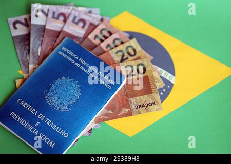 Tessera di lavoro brasiliana, libro blu di sicurezza sociale e banconote in denaro reais sulla bandiera della Repubblica Federativa del Brasile primo piano. Scheda di lavoro brasiliana e. Foto Stock