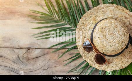 Un cappello di paglia si trova accanto a un paio di occhiali da sole su una superficie di legno, circondato da foglie di palma. Il cappello e gli occhiali da sole gettano un'ombra nella calda luce solare Foto Stock