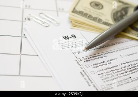 Modulo IRS W-9 richiesta di numero di identificazione contribuente e certificato vuoto si trova con la penna e molte banconote da cento dollari sulla pagina del calendario. Tassa Foto Stock