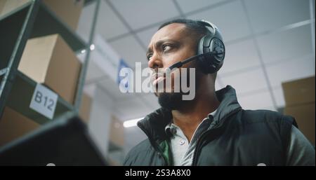 Coordinatore logistico afroamericano, responsabile che indossa le cuffie al cliente, utilizza un tablet computer nella struttura di magazzino. Centro di distribuzione logistica con ordini online di e-Commerce. Primo piano. Foto Stock