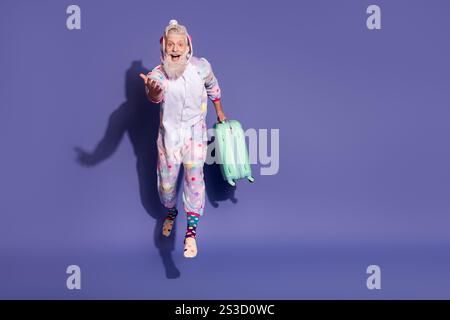 Gioioso uomo barbuto in pigiama unicorno con valigia su sfondo viola Foto Stock