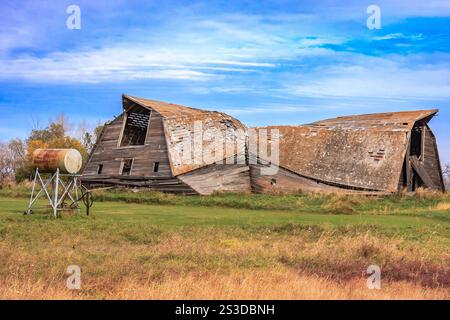 Un grosso, vecchio fienile con una vasca sopra. Il fienile è in un campo con erba e alberi sullo sfondo. Il fienile sembra essere in stato di abbandono Foto Stock