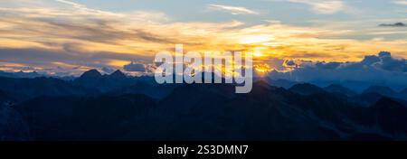 La luce del sole dorata proietta un caldo bagliore sulle aspre cime di una catena montuosa al crepuscolo. Le nuvole strisciano nel cielo mentre il giorno passa nella notte, creando una vista mozzafiato. Foto Stock