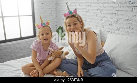 Madre e figlia fanno una videochiamata, salutano la telecamera mentre si siedono su un letto in un'accogliente camera da letto con un muro di mattoni bianchi e una grande finestra. Foto Stock