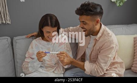 Coppia che celebra con entusiasmo il test di gravidanza positivo in un salotto accogliente con donna che tiene il test e uomo sorridente Foto Stock