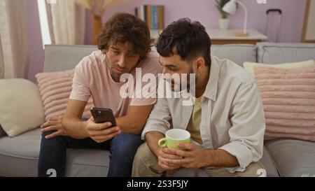 Due uomini seduti su un divano in un accogliente soggiorno, che guardano uno smartphone insieme, uno con una tazza verde e vari elementi di arredamento in casa Foto Stock