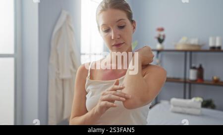 Donna bionda con scottature che applica lozione rilassante all'interno in un ambiente spa con asciugamani e prodotti benessere sullo sfondo Foto Stock