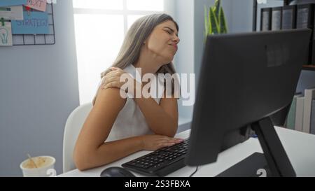 Giovane donna che soffre di dolore alle spalle in un ufficio, seduta a un computer con un'espressione dolorosa, evidenziando le sfide del prolungamento Foto Stock