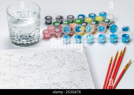 Kit di pittura per numeri con vasi, pennelli, tela e un bicchiere d'acqua su sfondo bianco. Divertimento creativo. Foto Stock