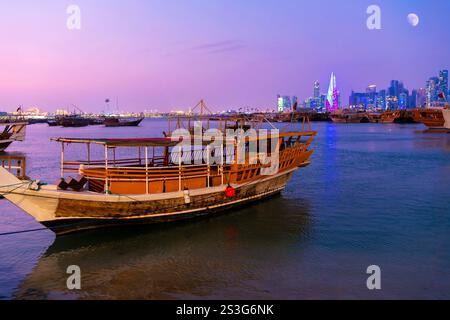 Barche tradizionali in dhow lungo la Corniche sul lungomare con lo skyline illuminato in lontananza all'ora blu crepuscolo, a Doha in Qatar. Foto Stock