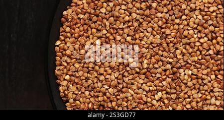 Grano saraceno crudo in un piatto nero su un tavolo di legno scuro. Semole di grano saraceno in un piatto, vista dall'alto. Grano saraceno crudo, porridge crudo. Foto Stock