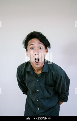 Un giovane uomo in camicia nera con un'espressione scioccata, occhi larghi e bocca aperta, appoggiato leggermente in avanti su un semplice sfondo bianco, capturin Foto Stock