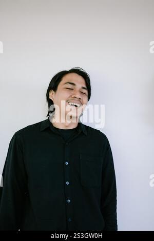 Un giovane allegro con una camicia nera che ride gioiosamente su un semplice sfondo bianco, catturando un momento schietto e spensierato. Foto Stock