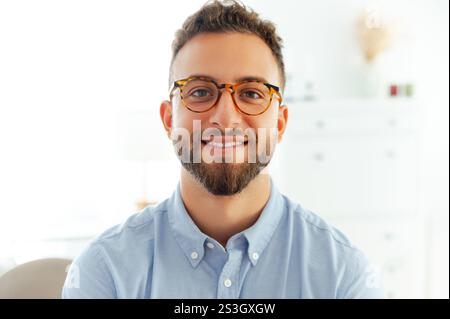 Ritratto ravvicinato di un uomo sorridente e di successo con occhiali e camicia blu, aspetto sicuro e professionale. Attraente, positivo uomo caucasico o arabo stare in ufficio, guardando la fotocamera Foto Stock