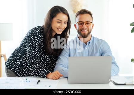 Concetto di lavoro di squadra. Donna sorridente in una camicia appoggiata su un uomo con una camicia blu, che guarda entrambi lo schermo di un notebook e collabora a un progetto in un ambiente luminoso e moderno con grafici sul tavolo Foto Stock