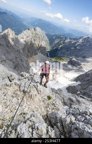 Alpinista con casco in ferrata, Zugspitze via ferrata, salita allo Zugspitze, vista del bacino montano con campo innevato della Hoelle Foto Stock