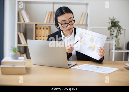 Una donna d'affari asiatica che indossa le cuffie si occupa di videochiamate online mentre esamina i documenti alla moderna scrivania dell'ufficio. Foto Stock