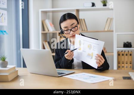 Una donna d'affari asiatica che indossa le cuffie si occupa di videochiamate online mentre esamina i documenti alla moderna scrivania dell'ufficio. Foto Stock