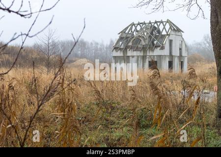 Una casa abbandonata, parzialmente costruita, sorge tra erba alta in una giornata nebbiosa, circondata da un paesaggio desolato e sovrastato. Foto Stock