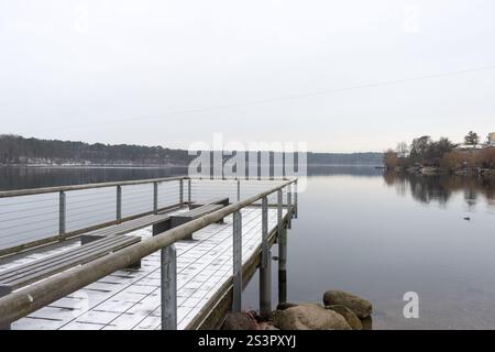 Una serena vista invernale di un molo innevato che si estende su un lago calmo e riflettente circondato da un paesaggio tranquillo. Foto Stock