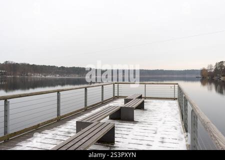 Un tranquillo scenario invernale caratterizzato da un molo in legno con panche coperte di neve, che si affaccia su un tranquillo lago ghiacciato circondato da foreste. Foto Stock