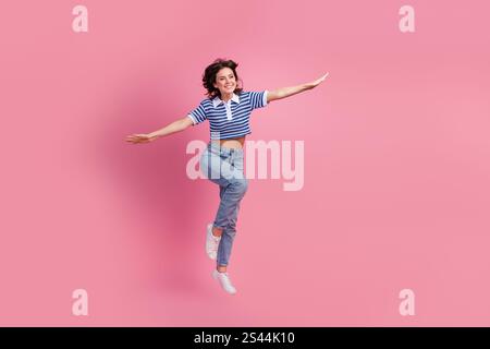 Una donna allegra con t-shirt a righe e jeans salta gioiosamente su uno sfondo rosa vibrante, incarnando energia e felicità giovanile Foto Stock