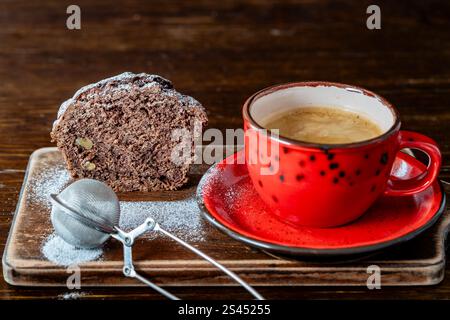 Chocolate muffin with red cherries and walnuts sprinkled with powdered sugar and red cup of black coffee on a wooden table, close up. Homemade delicio Foto Stock