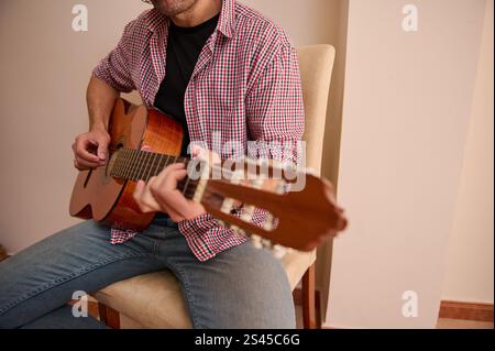 Un uomo che suona una chitarra acustica mentre è seduto all'interno, indossa una camicia a scacchi e un jeans casual. La messa a fuoco mette in risalto la mano e lo strumento, il cappuccio Foto Stock