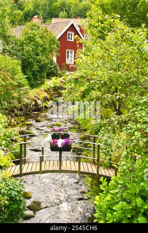 Ruscello con un piccolo ponte di fronte a una casa di legno rossa in Svezia. Alberi in banca . Natura scandinava Foto Stock