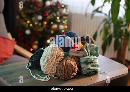Una collezione di palline colorate in filato e forniture per maglieria disposte in modo ordinato su un tavolo, disposte su uno sfondo accogliente con luci festose e una festa natalizia Foto Stock