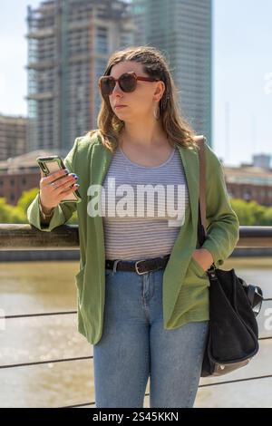 Una giovane donna che indossa occhiali da sole, un top a righe e una giacca verde, guarda il suo telefono mentre si trova vicino a un porto, con edifici alti e un fiume Foto Stock