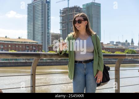 Una giovane donna sorridente che indossa occhiali da sole, una parte superiore a righe e una giacca verde, tenendo il telefono vicino a un fiume, con moderni edifici cittadini sullo sfondo Foto Stock