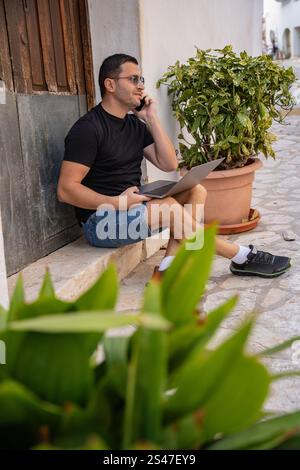 Uomo calmo che parla al telefono cellulare usando un computer portatile all'aperto per strada nella città vecchia spagnola di Guadalest Foto Stock
