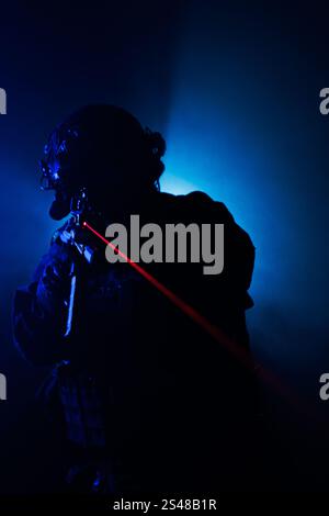 Una figura tattica sagomata che regge un'arma dotata di un laser rosso, illuminato da un'illuminazione blu scuro, che trasmette mistero e intensità in un dondolo Foto Stock