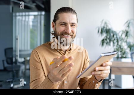 Persona con la barba che indossa un abbigliamento casual prende appunti con entusiasmo in un ufficio professionale, suggerendo la collaborazione. Foto Stock