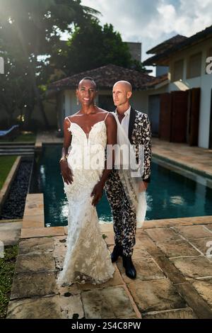 Persona fluida di genere in abito bianco con sposo LGBTQ in tuta vicino alla piscina al matrimonio tropicale. Celebra l'amore e la diversità in un ambiente esotico. Perfetto per Foto Stock