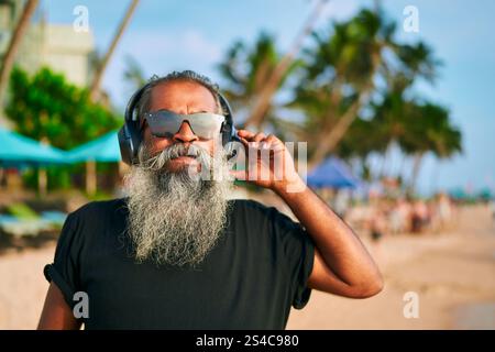 L'uomo anziano con la barba grigia ama la musica sulla spiaggia tropicale. L'elegante nonno indossa occhiali da sole e ascolta le cuffie moderne. Atmosfera rilassante e piacevole Foto Stock