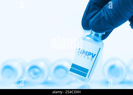 Una mano blu dei medici con i guanti che tiene in mano una fiala di vaccino etichettata HMPV virus, con una fila sfocata di fiale di vaccino sullo sfondo. Foto Stock
