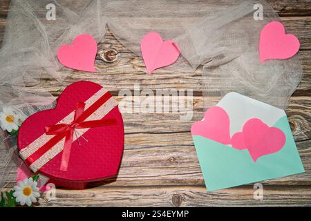 scatola regalo a forma di cuore con cuori di carta, busta e fiori su un tavolo di legno Foto Stock