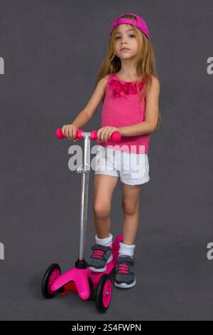 Una bambina di quattro anni indossa uno stile sportivo e tiene in mano uno scooter per bambini. Fotografato su sfondo bianco Foto Stock