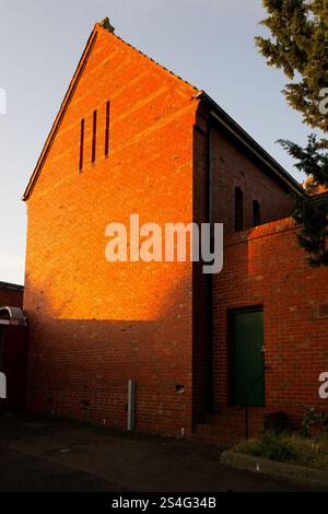 Parte posteriore di un edificio chiesa in mattoni rossi illuminato dal tramonto, con cortile pavimentato e porta verde in legno all'ombra Foto Stock