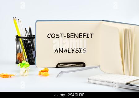 Un notebook aperto sulla pagina denominata analisi costi/benefici si trova su una scrivania accanto alla cancelleria, indicando i preparativi per una valutazione aziendale o. Foto Stock