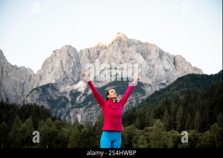 Una donna con giacca rosa e leggings blu alza con gioia le braccia, in piedi su uno sfondo mozzafiato di montagne aspre e foreste lussureggianti. Foto Stock